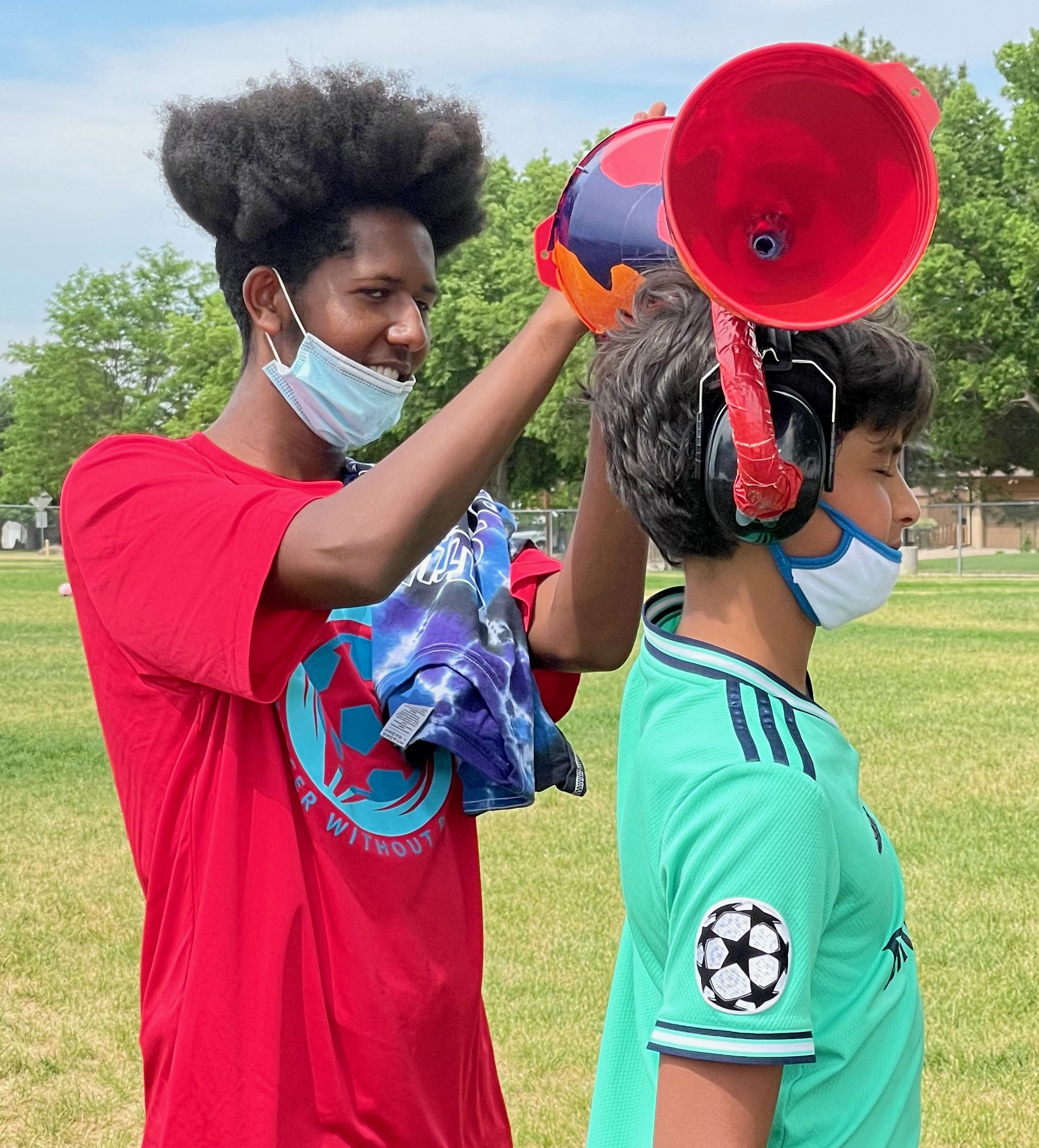 High school aged student is clapping their hands near a middle school student who is wearing hearing protect earmuffs that have been modified with tubing and funnels. This configuration maps sounds on your left side to your right ear and vice versa.