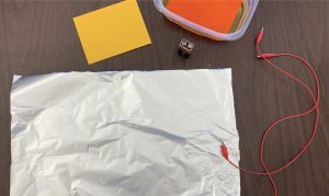 There is sheet of aluminum foil, wires, a 9 Volt battery, a yellow piece of paper, and a reddish piece of paper in water in a plastic dish.