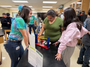 women in tie dye shirt on left of photo shows two high school age students in right of photo an experiment with small clothing on a wire